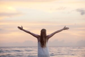 Person standing by the ocean with arms wide open at sunrise, symbolizing freedom and overcoming fear.