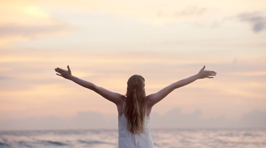 Person standing by the ocean with arms wide open at sunrise, symbolizing freedom and overcoming fear.